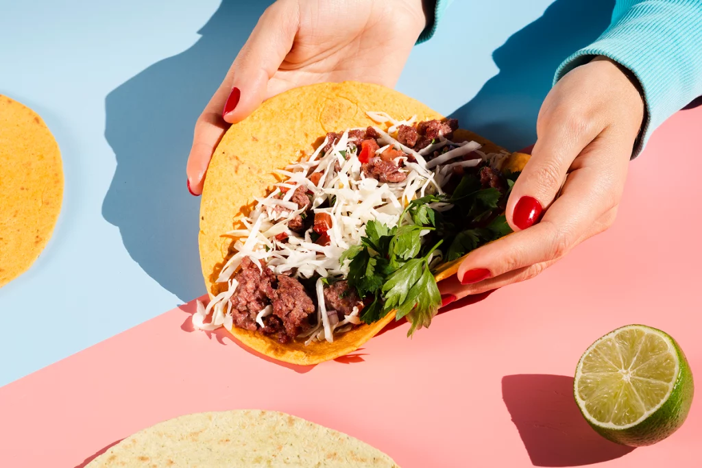 Taco garni de bœuf, fromage râpé et coriandre tenu entre deux mains.